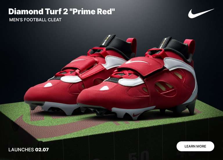 Pair of red, white, and black Nike Diamond Turf 2 Prime Red men’s football cleats displayed on a turf-covered pedestal with launch date and Nike branding.