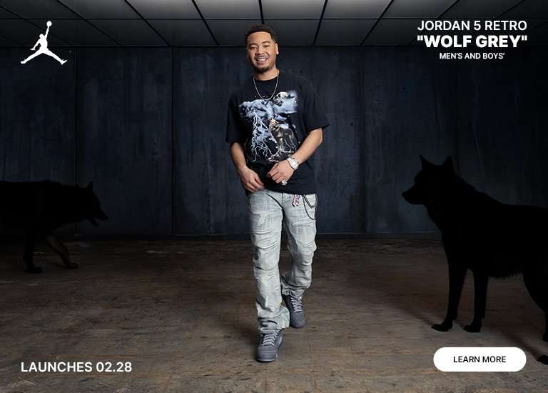An advertisement for Jordan 5 Retro Wolf Grey sneakers. A man wears the shoes in a dark, industrial setting with wolf silhouettes in the background. A close-up of the grey suede sneaker highlights the 23 branding. Text states the shoes for men and boys launch 02.28 with a Learn More button.
