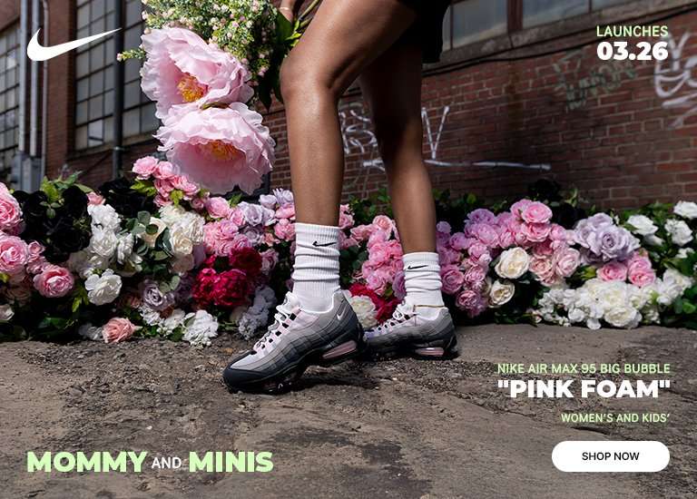 Promotional banner for Nike Air Max 95 Big Bubble Pink Foam featuring a woman with blonde hair and a child wearing matching sneakers. The scene is set outdoors against a brick wall and a bed of pink flowers. Text on the image includes Mommy and Minis, Launches 03.26, and a Shop Now button.
