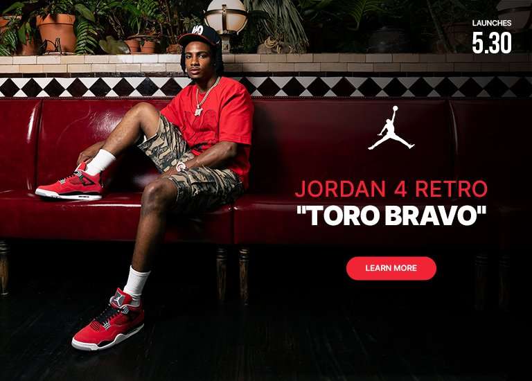 A promotional banner featuring a man sitting on a long red leather booth, modeling red Jordan sneakers, a red t-shirt, and camouflage shorts. The setting has an upscale, moody vibe with plants and lighting fixtures along the back wall. On the right side, the white Jordan Jumpman logo sits above the text JORDAN 4 RETRO TORO BRAVO. In the top right corner, text reads LAUNCHES 5.30, and a red LEARN MORE button is at the bottom right.