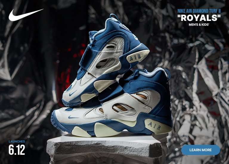 A Nike promotional banner features two Nike Air Diamond Turf II Royals sneakers stacked on a white stone block against a crumpled, metallic silver background. The mid-top sneakers showcase a white and deep blue upper with a midfoot strap and a gold mesh underlay panel. A white Nike swoosh is in the top left, blue text in the bottom left reads LAUNCHES 6.12, and the top right text reads NIKE AIR DIAMOND TURF II ROYALS MEN'S & KIDS'. A blue LEARN MORE button sits in the bottom right corner.