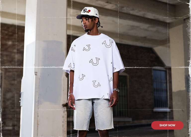 Lifestyle banner featuring a male model in a True Religion outfit. He is wearing a white oversized t-shirt with black horseshoe logos, light-wash denim shorts, and a New York Yankees hat with a red rose detail. The background shows an industrial urban setting with a red Shop Now button in the bottom right corner.