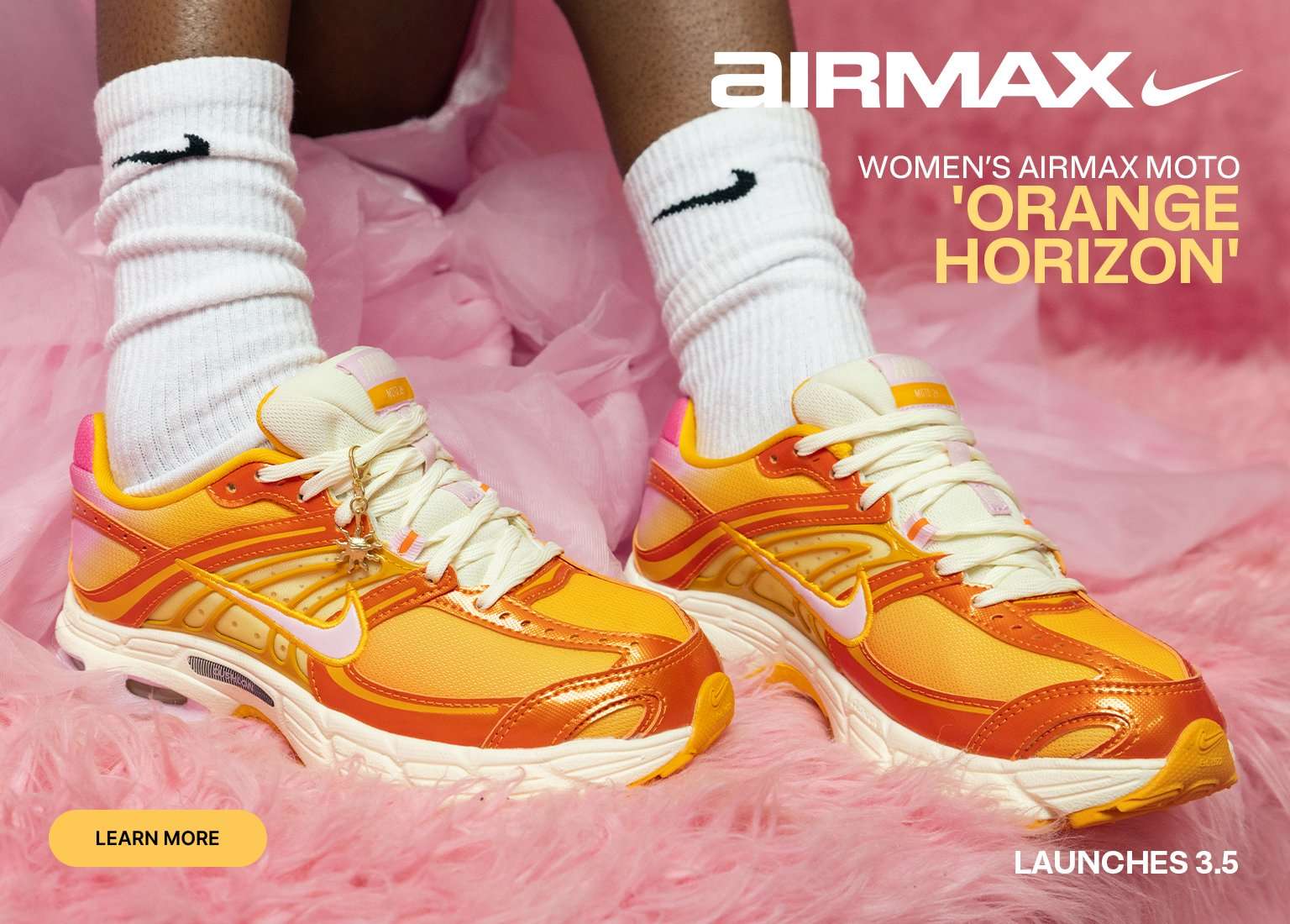 Promotional banner for the Women's Airmax Moto Orange Horizon. The image features a close-up of the sneakers, which have a vibrant orange, yellow, and pink gradient design with cream-colored laces and midsoles. A small gold sun charm is attached to the laces of the left shoe. The shoes are set against a fluffy pink background and are being worn with white Nike socks. The Air Max logo is in the top left, and the text indicates a launch date of 3.5.