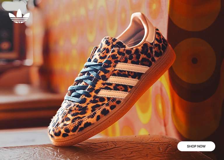 An adidas Handball Spezial sneaker featuring a cheetah print pony hair upper, white three-stripes, and blue laces. The shoe is angled upward against a retro, warm-toned patterned background with an adidas logo in the top left and a SHOP NOW button at the bottom right.