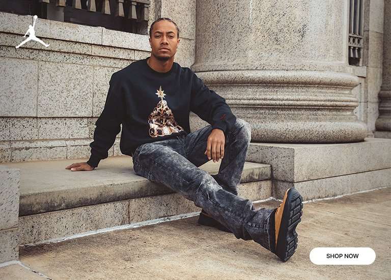 A man sits on stone steps in front of a large classical column, wearing a navy crewneck sweatshirt featuring a graphic of a Christmas tree made of sneakers. He is wearing acid-wash jeans and tan Air Jordan City Boots with thick black soles. A white Jordan logo is in the top left corner, and a SHOP NOW button is in the bottom right.