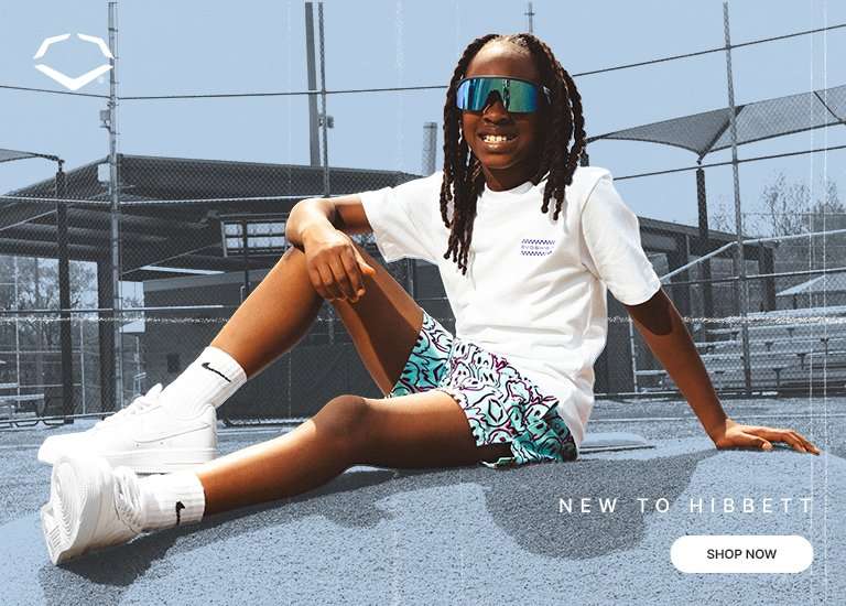 A young boy wearing blue sunglasses sits on a baseball field mound, wearing a white EvoShield t-shirt, patterned shorts, and white Nike Air Force 1 sneakers. The banner features an EvoShield logo and New to Hibbett text with a Shop Now button.