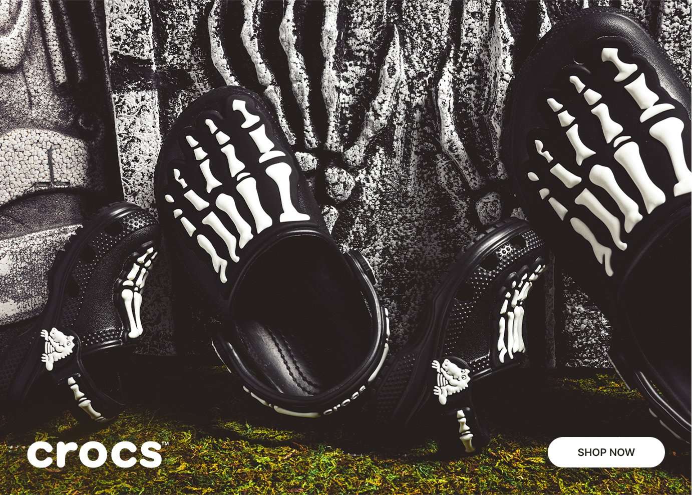Black Crocs clogs and sandals featuring glow-in-the-dark skeleton bone designs displayed on a spooky textured backdrop with moss accents.