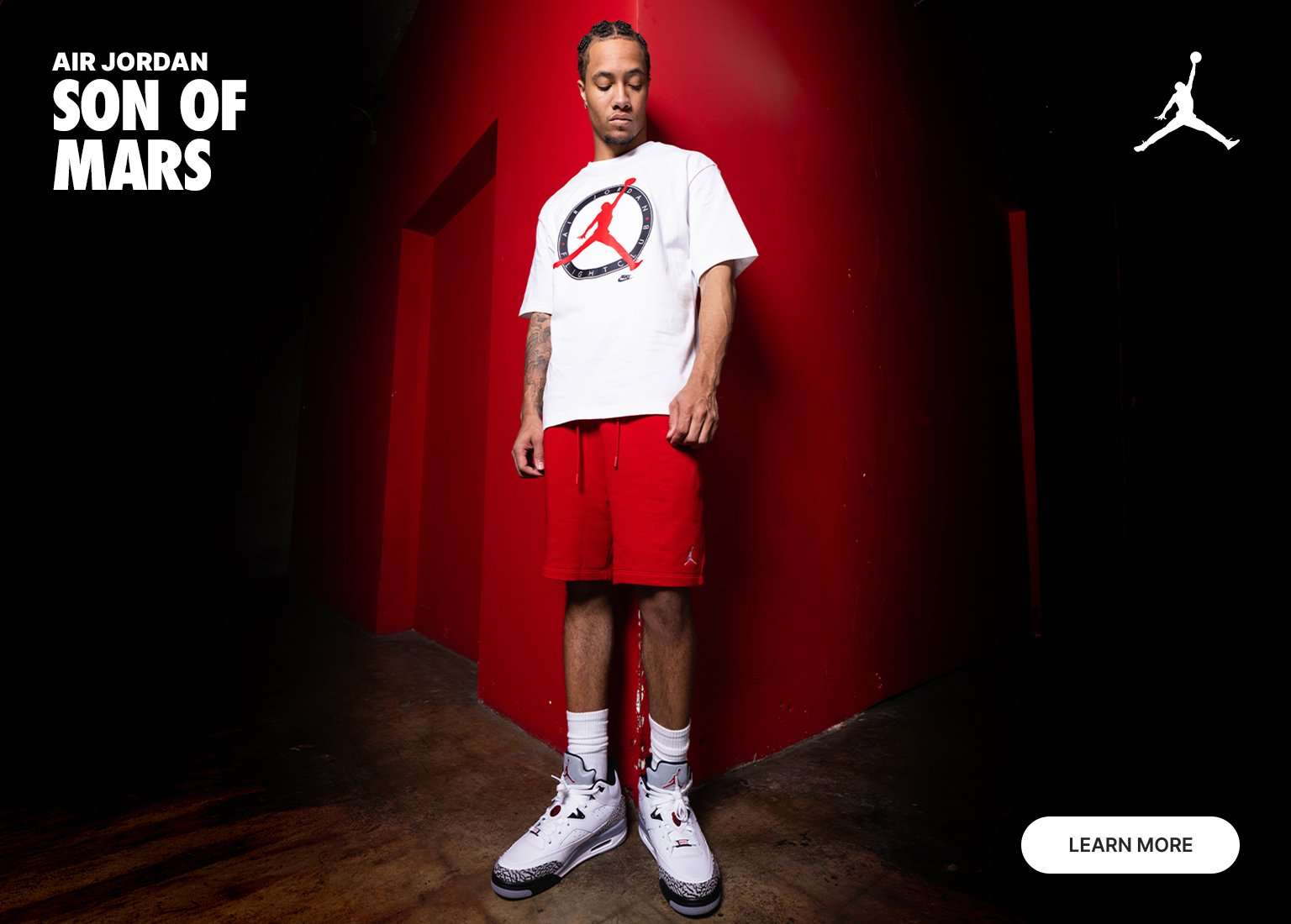A man stands in the corner of a red-walled room wearing a white Jordan Flight Club t-shirt, red shorts, and white and grey Air Jordan Son of Mars sneakers. The top left features the text AIR JORDAN SON OF MARS, while a white Jumpman logo is in the top right corner and a LEARN MORE button is in the bottom right.