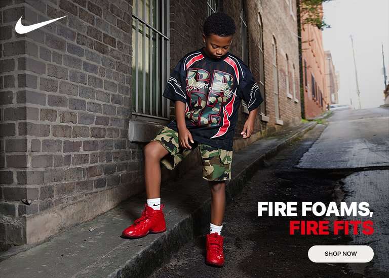 Boy adjusting bright red Nike Air Foamposite Pro Gym Red sneakers while wearing streetwear apparel in an urban setting. Promotion highlights the Nike Air Foamposite Pro and coordinating youth apparel with a Shop Now call to action.
