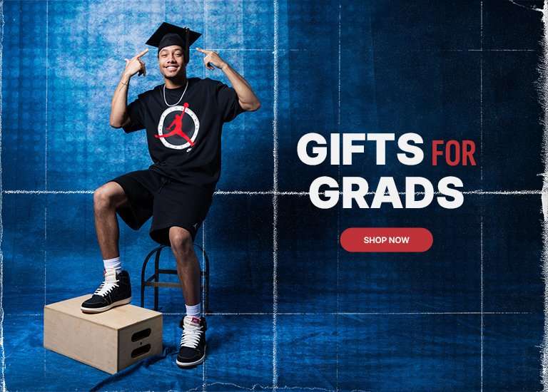 A promotional banner titled GIFTS FOR GRADS features a smiling young man in a graduation cap and black Jordan Brand apparel. He sits against a blue textured background, wearing black and white high-top sneakers with one foot resting on a wooden crate. A red SHOP NOW button is centered on the right.