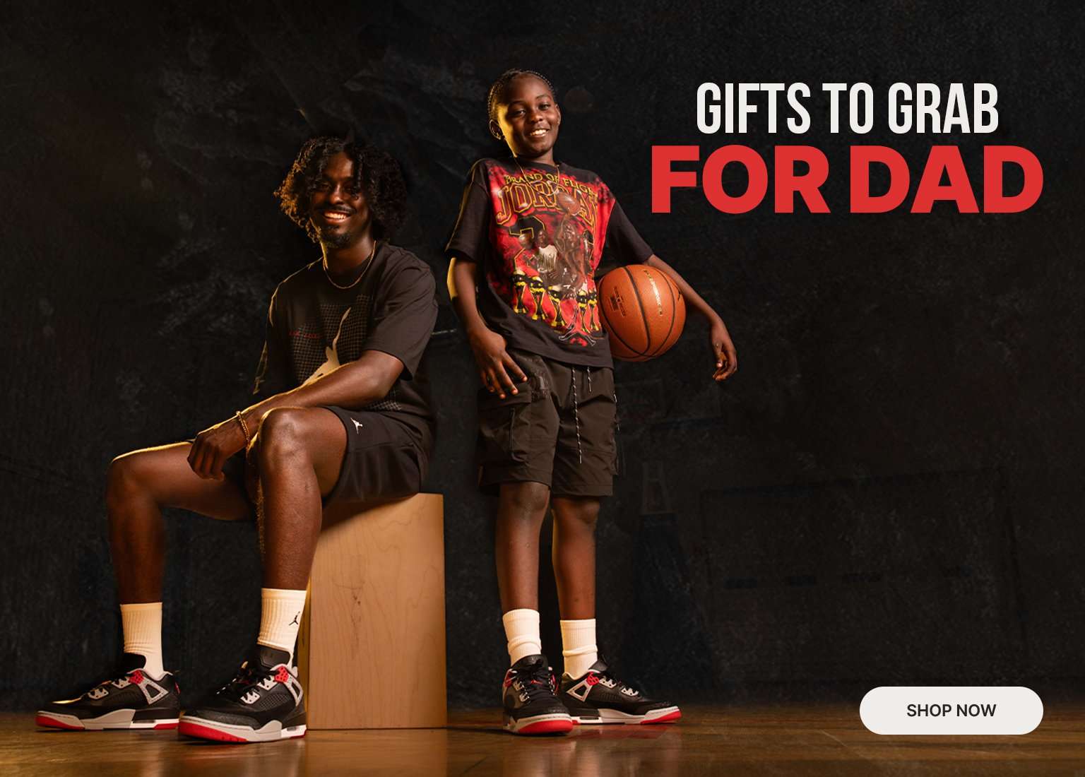 A marketing banner set against a dark textured backdrop featuring a man sitting on a wooden block and a smiling child standing beside him holding a basketball, both wearing matching black, red, and grey Jordan sneakers. On the right, bold text reads Gifts to Grab for Dad next to a white Shop Now button in the bottom corner.