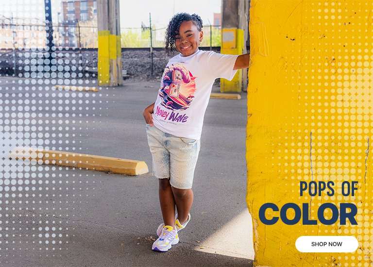A young girl smiling and leaning against a yellow pillar, wearing a graphic tee and New Balance 9060 sneakers in a vibrant lilac, yellow, and blue colorway. A large, detailed close-up of the New Balance 9060 sneaker is featured on the left, highlighting its chunky silhouette and multi-colored suede overlays. The background has a blue halftone dot pattern and the text New Balance 9060, Pops of Color, and a Shop Now button.