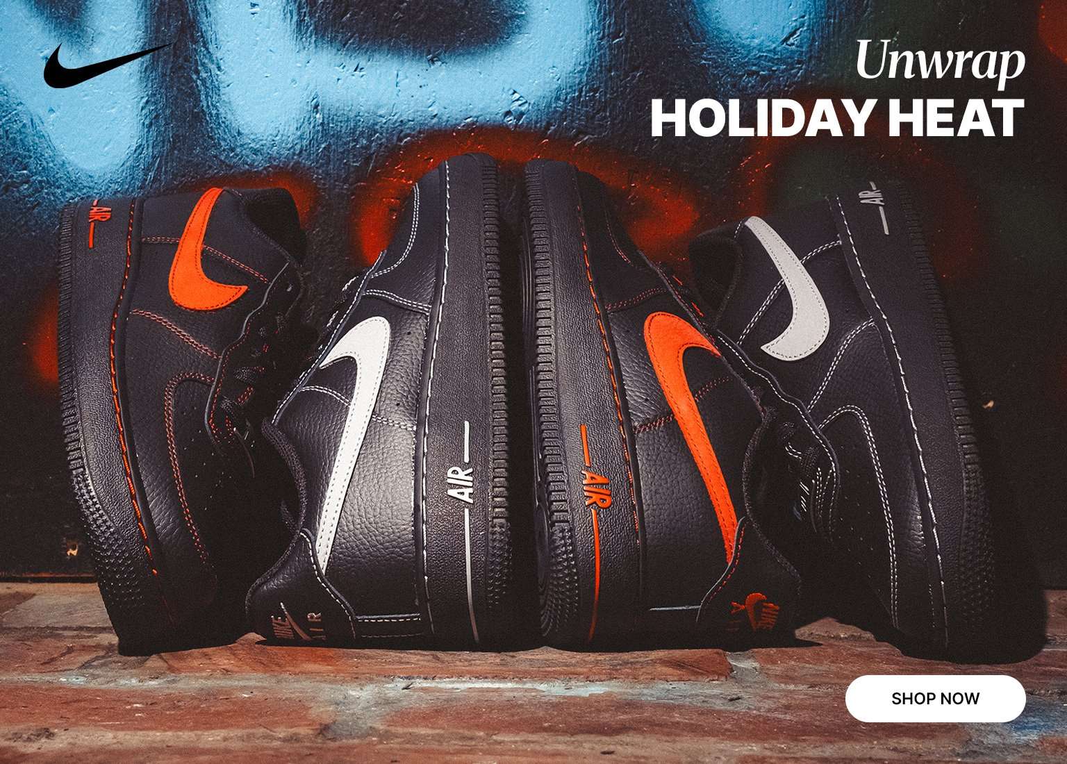 An array of black Nike Air Force 1 LV8 sneakers, featuring white or orange Swooshes, are displayed against a dark blue background with graffiti. The shoes are standing upright on a brick surface, and the text below reads, Unwrap HOLIDAY HEAT, advertising them as a seasonal gift.