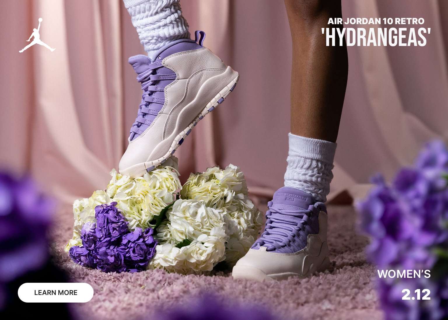 A promotional banner for the women’s Air Jordan 10 Retro 'Hydrangeas' featuring the sneakers styled with thick white slouch socks amidst a bed of purple and white hydrangeas. The shoes showcase a soft pastel color palette of off-white leather and lavender accents, with a release date of February 12th displayed in the corner.