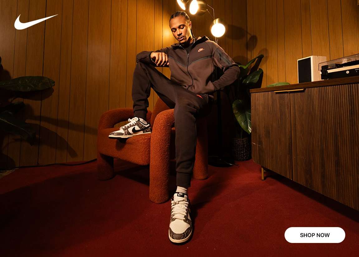 Nike athlete in brown tracksuit and black-and-white sneakers seated in a retro lounge setting.