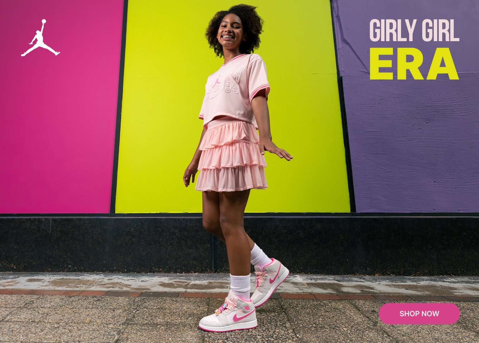 A promotional banner features a young girl posing and walking in a pink Jordan crop top, layered ruffle skirt, and white Air Jordan 1 Mid sneakers accented with pink and floral lace charms. The background is a bright neon yellow wall with a pink border on the left showcasing a white Jordan Jumpman logo. Center text reads GIRLY GIRL ERA, and a pink SHOP NOW button sits in the bottom right corner.