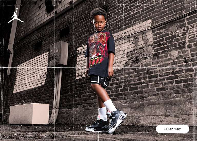 A young boy posing in an alley wearing a Jordan graphic t-shirt, athletic shorts, and black cement Jordan sneakers. A white Jordan Jumpman logo is in the top left, and a Shop Now button is in the bottom right.