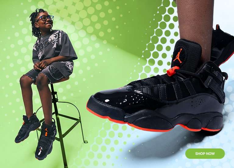 A young boy sits on a green stool wearing a grey graphic tee, denim shorts, and black and red Jordan 6 Rings sneakers. A large, detailed close-up of the sneaker is featured in the foreground against a green and blue halftone dot background with a Shop Now button in the corner.