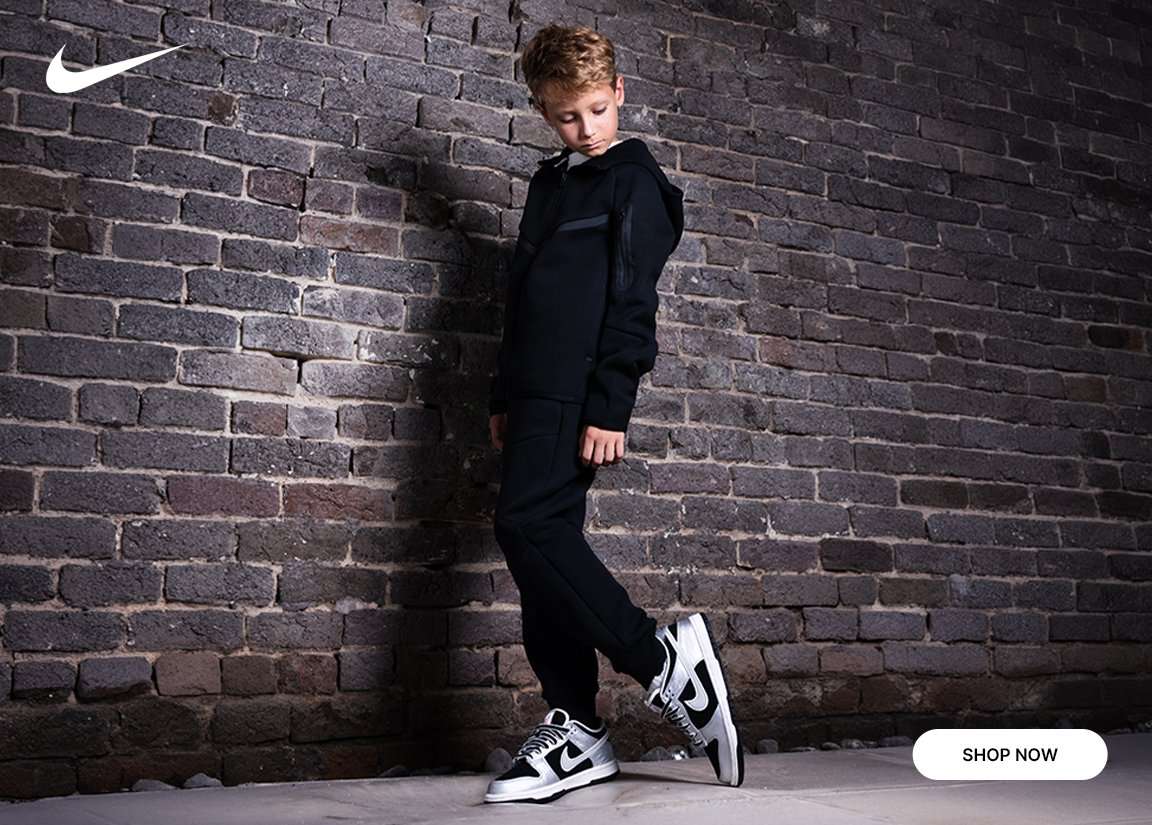 Kids model wearing a black Nike Tech Fleece set with black and silver Nike Dunk Low sneakers against a brick wall backdrop.