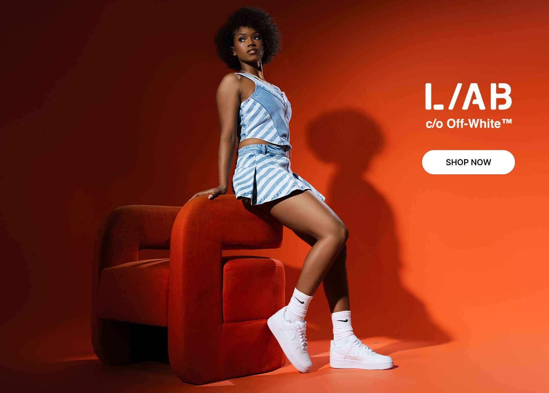 L.A.B c/o Off-White denim top and pleated skirt set styled with white Nike sneakers against a red studio backdrop.
