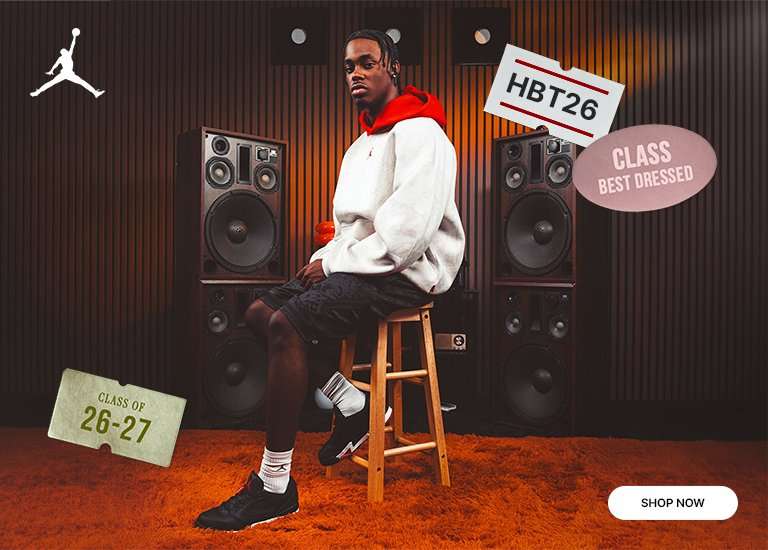 Jordan Air 1 sneaker in black with red stitching displayed on an orange shag rug alongside a coordinated Jordan fleece outfit. The scene features back-to-school themed graphics, speakers, and a seated model wearing the collection, with a Shop Now button.