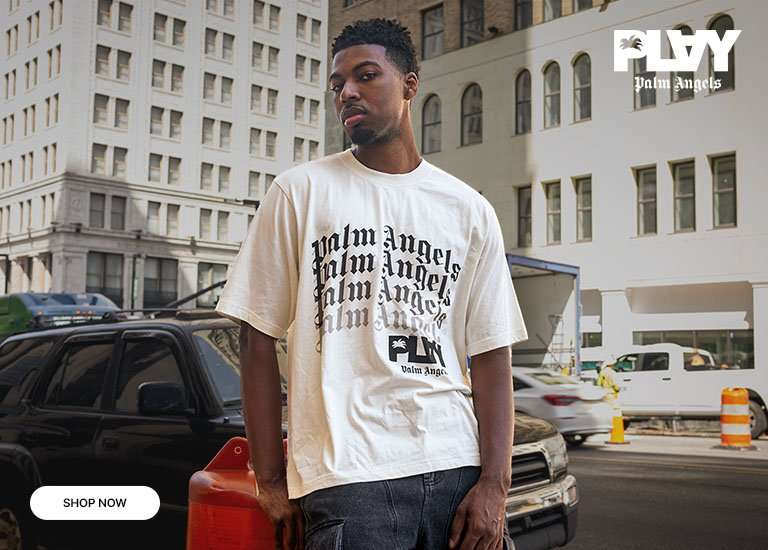 Palm Angels graphic T-shirt in cream featuring stacked logo text, worn in an urban city setting with buildings and parked vehicles in the background.