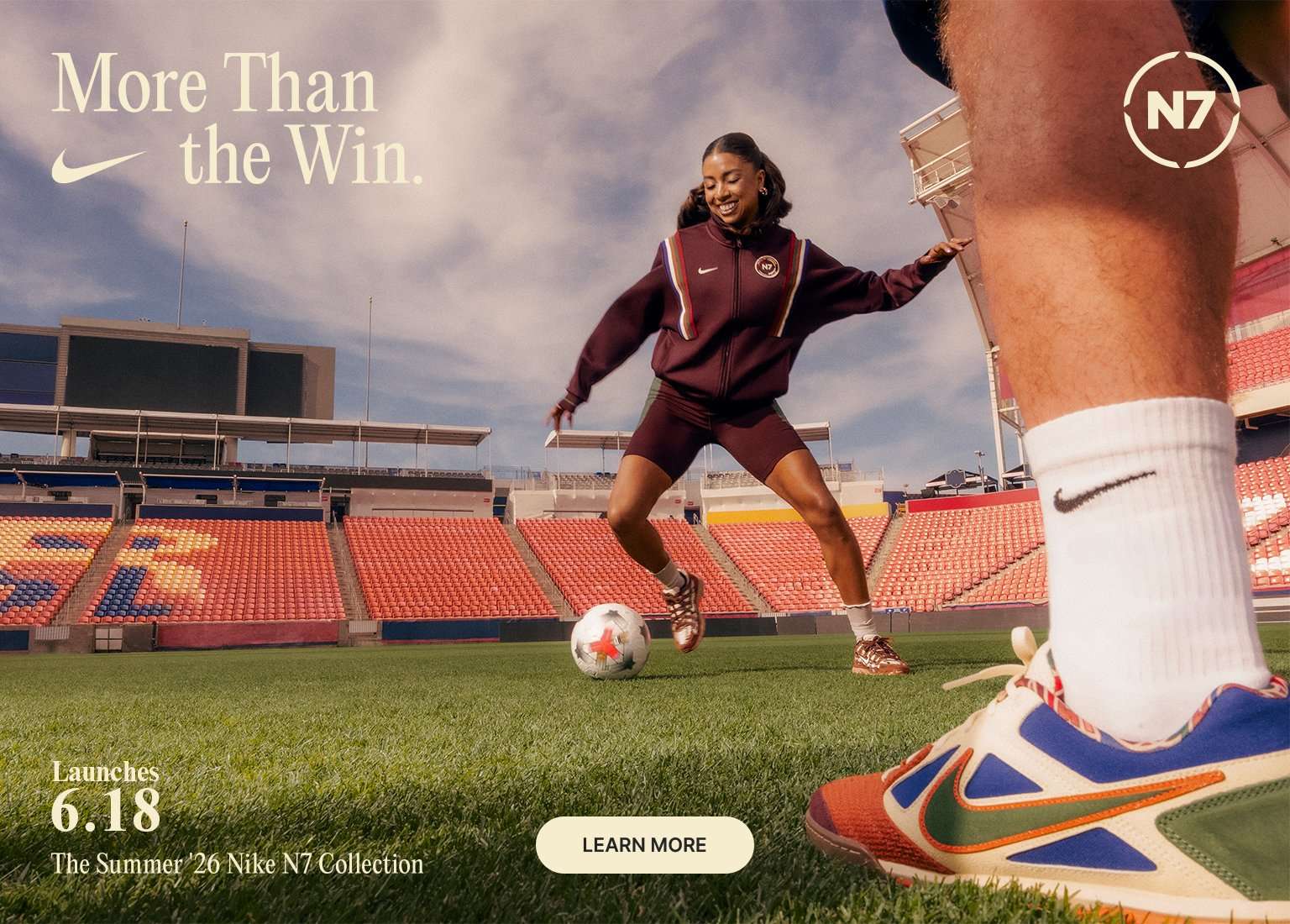 A woman dribbles a soccer ball on a stadium field wearing a Nike N7 tracksuit. A close-up in the foreground shows a player's colorful Nike sneaker. Promotional text reads, More Than the Win. Launches 6.18. The Summer '26 Nike N7 Collection.