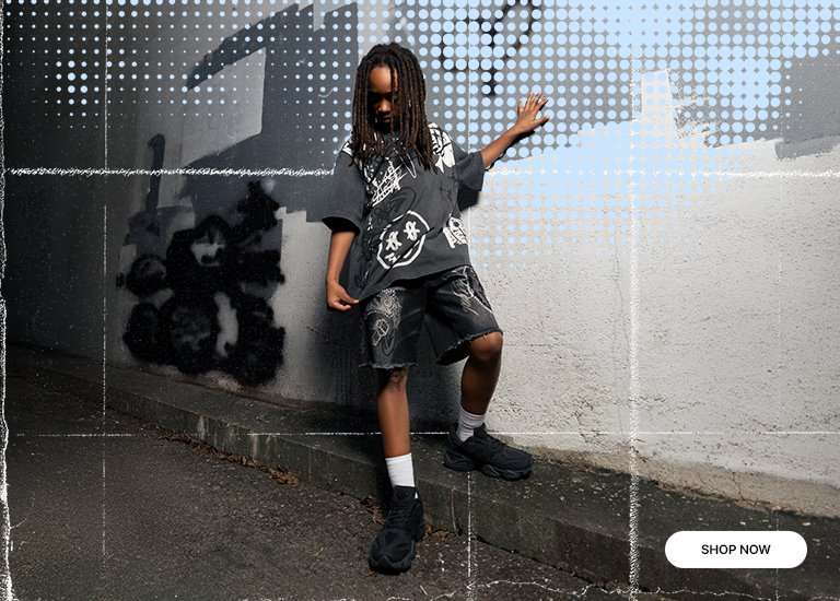 A promotional banner for New Balance featuring triple black 9060 sneakers. On the left, a young boy with locs poses in a graphic tee and denim shorts against a graffiti-covered wall with a heart. The right side shows a close-up of the sneakers and white socks. The design includes a blue halftone dot overlay and a Shop Now button in the bottom right corner.