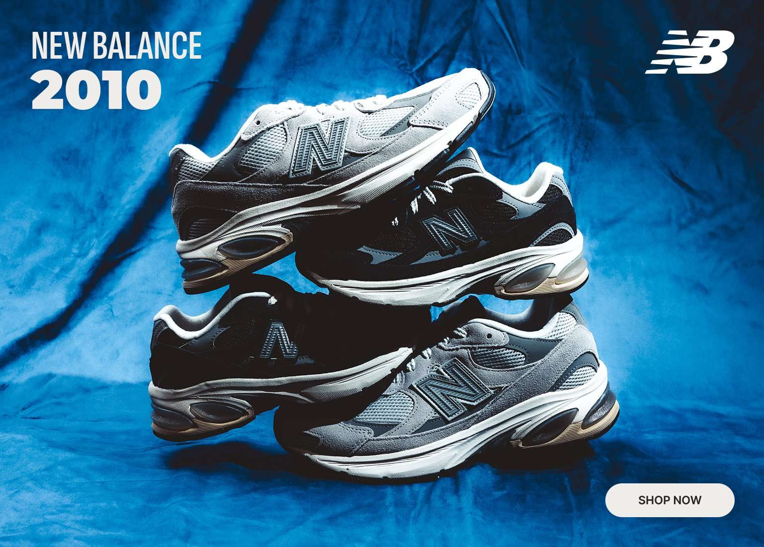 A stack of four New Balance 2010 sneakers in grey and black colorways is centered against a draped blue fabric background. The top left corner features the text NEW BALANCE 2010, the top right displays the white NB logo, and a white SHOP NOW button is located in the bottom right.
