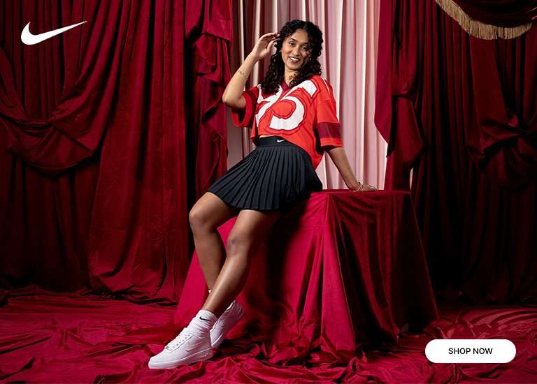 A woman posing in a red cropped jersey, black pleated skirt, and white Nike Air Force 1 sneakers against a backdrop of draped red velvet curtains.