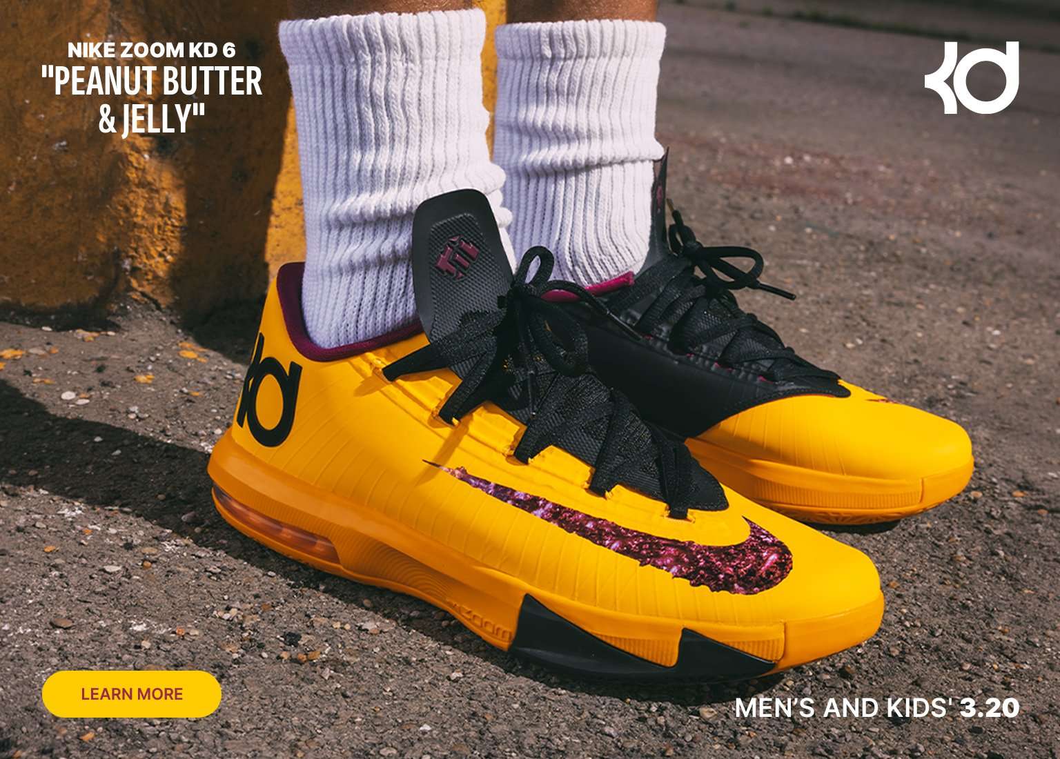 A person wears the Nike Zoom KD 6 Peanut Butter & Jelly sneakers with white crew socks. The shoes feature a bright yellow upper, black tongue and laces, and a maroon jelly-textured Swoosh. The background shows a paved surface and a yellow concrete pillar, with a Learn More button and 3.20 release date text overlaid.