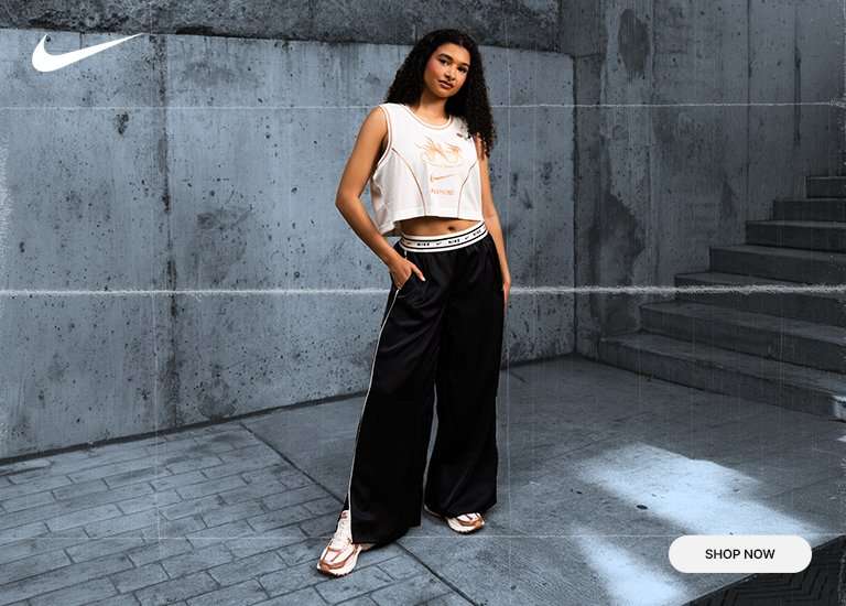 A promotional banner for Nike featuring a close-up of a Nike Zoom Vomero 5 sneaker in cream and rose gold. To the right, a woman models a matching white crop top and black wide-leg trousers in an urban concrete setting. A Shop Now button is in the bottom right corner.