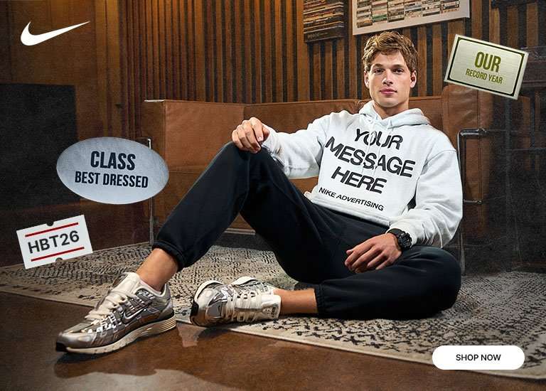 Nike model wearing a white hoodie, black joggers, and silver sneakers while seated indoors with back-to-school themed graphics and Nike branding.