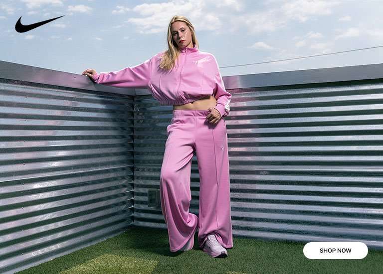Person wearing a pink Nike track jacket and wide-leg track pants set while posing outdoors on a metal rooftop, paired with pink and white Nike sneakers.
