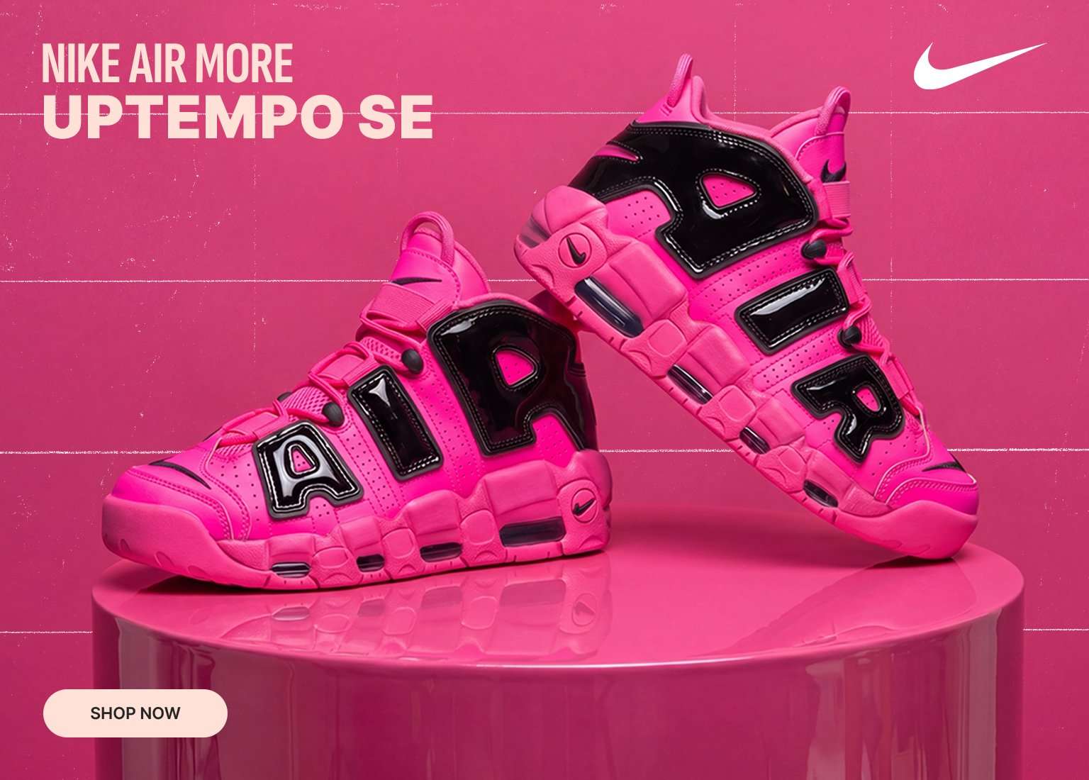 A pair of bright pink Nike Air More Uptempo SE sneakers with glossy black AIR lettering displayed on a pink pedestal against a matching pink background. The Nike logo and a Shop Now button are visible.