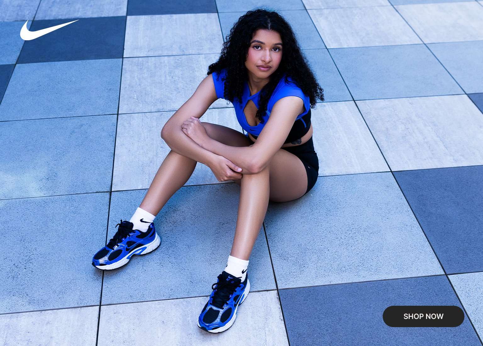 Lifestyle banner featuring a model in a blue and black Nike performance outfit and biker shorts, seated on a checkered pavement. The image includes a close-up crop of blue, black, and white Nike sneakers with white crew socks. A Shop Now button and white Nike Swoosh are visible on the layout.