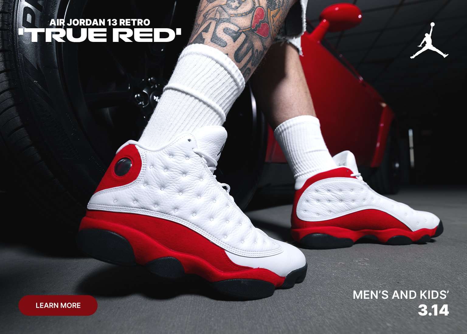 A low-angle shot of a person wearing white and red Air Jordan 13 Retro True Red sneakers, standing next to a black tire and a red sports car. The graphic includes the Jumpman logo and text indicating Men’s and Kids’ sizes with a 3.14 release date.