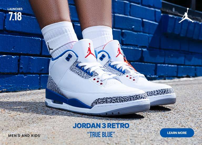 Close-up of the Air Jordan 3 Retro True Blue sneakers in white, blue, and gray with elephant print details, displayed outside a bright blue brick building. A person wearing the sneakers stands in the background, highlighting the July 18 launch for men’s and kids’ sizes.