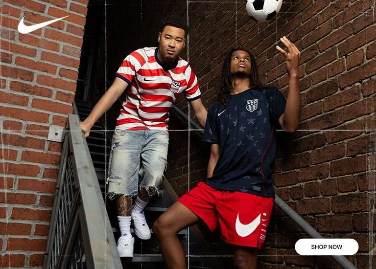A wide banner featuring two men modeling Nike U.S. Soccer jerseys in urban settings like stairs and brick alleyways. One man wears a red and white striped jersey, while the other wears a navy blue jersey patterned with stars. They pose with soccer balls and wear Nike sneakers. A white Nike swoosh is in the top left corner, and a SHOP NOW button is in the bottom right corner.