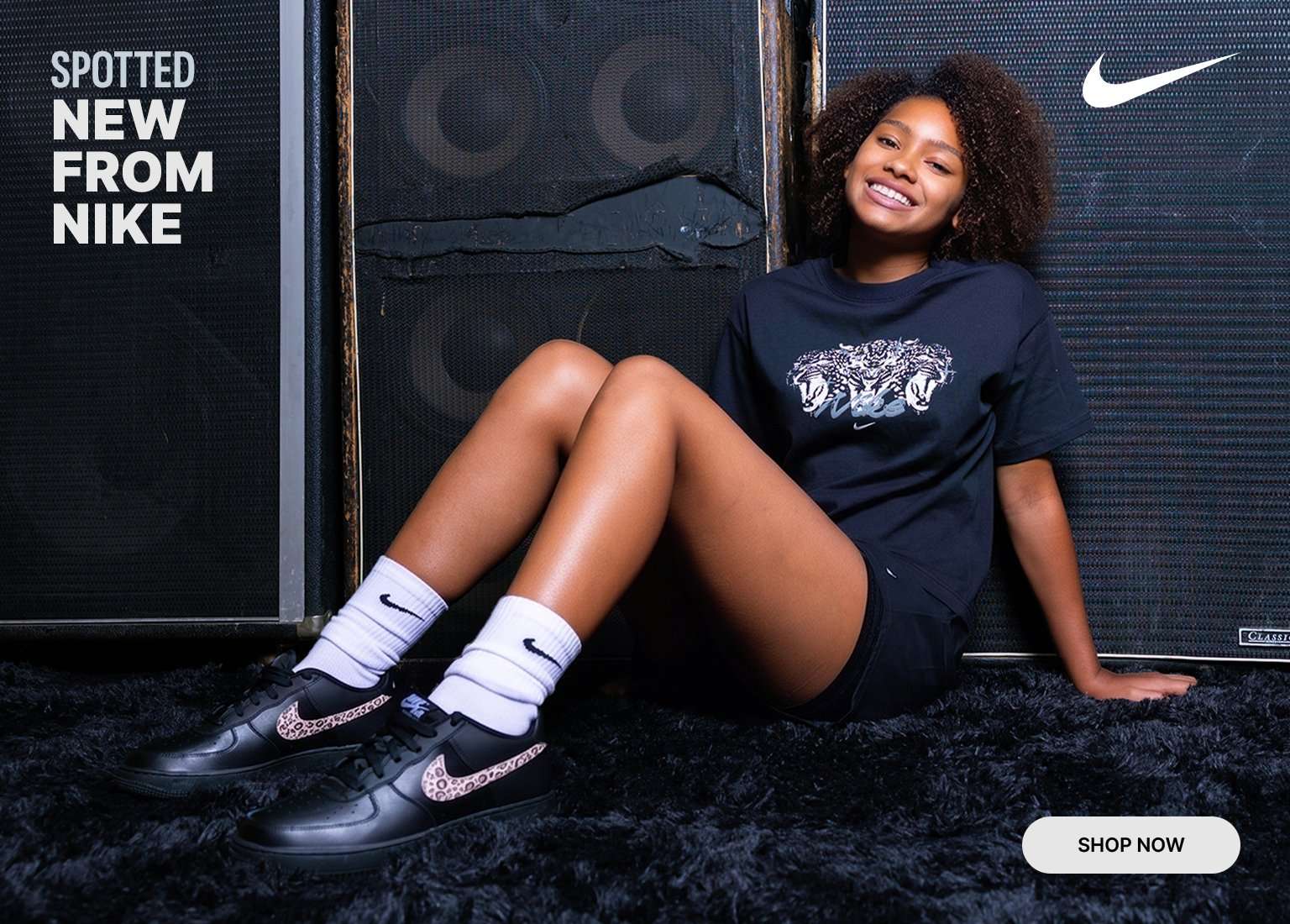 Person seated in front of large speaker cabinets wearing a Nike graphic tee, shorts, crew socks, and Nike sneakers, promoting new Nike apparel and footwear arrivals.