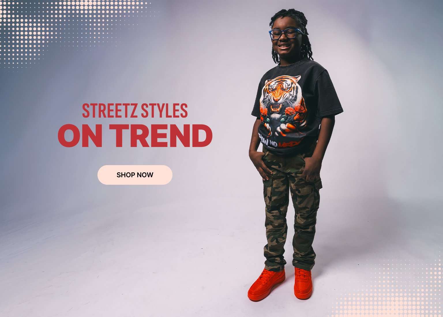An advertisement for Streetz Styles featuring a young person wearing a black t-shirt with a large tiger graphic, camouflage cargo pants, and bright orange sneakers. The left side of the image has a red textured background with white halftone dots and text that reads STREETZ STYLES ON TREND above a SHOP NOW button.