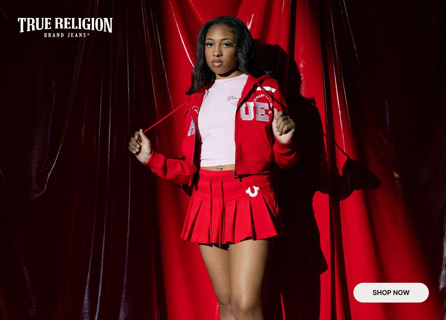 True Religion Brand Jeans featuring a model in a red zip-up hoodie with white lettering and a matching pleated mini skirt over a light pink tee. The layout includes a full-body shot and a close-up against a red draped backdrop with a Shop Now button.