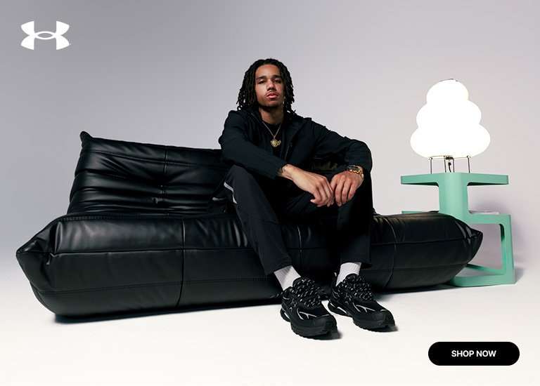 Under Armour athlete in an all-black outfit seated on a black leather sofa wearing black athletic sneakers.
