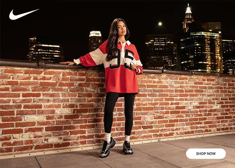 A woman with long dark hair poses on a rooftop at night against a city skyline. She is wearing an oversized red and white rugby polo with the number 95 on the front, paired with black leggings, white socks, and silver and black sneakers. A white Nike swoosh logo is visible in the top left corner.