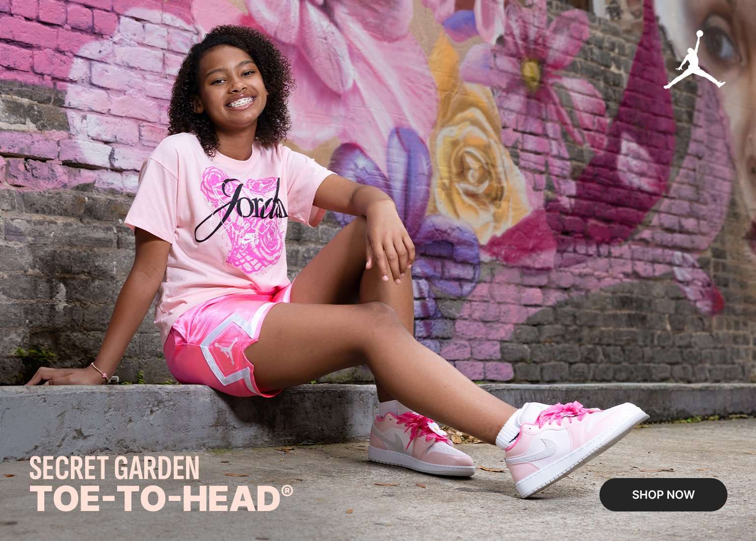 A promotional banner for the Jordan Secret Garden collection. The left side features a close-up of light pink Air Jordan 1 Low sneakers with bright pink laces and colorful flower charms. On the right, a young girl sits against a brick wall with a floral mural, wearing a matching pink Jordan T-shirt and shorts. The text SECRET GARDEN TOE-TO-HEAD appears in the bottom left, with a SHOP NOW button in the bottom right.