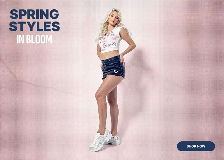 A model posing in a white graphic crop top, dark denim shorts, and white ASICS sneakers against a light pink gradient background. To the right, large navy and white text reads 'SPRING STYLES IN BLOOM' above a navy 'SHOP NOW' button.