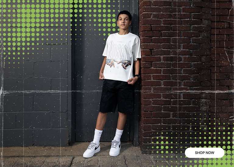 Professional streetwear lifestyle shot of a model leaning against a brick and charcoal wall. He is wearing a white Upscale graphic tee featuring a Creation of Adam inspired hand illustration, paired with black cargo shorts, white mid-calf socks, and white Nike Air Force 1 Lows with patterned laces. The image features a stylized green halftone dot overlay in the corners.