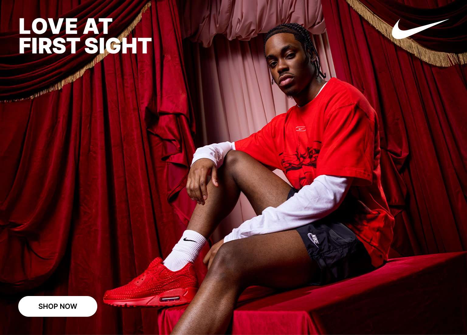 A man in red and black Nike apparel sitting on a stage with red velvet curtains, wearing all-red Nike Air Max sneakers, with text reading LOVE AT FIRST SIGHT.