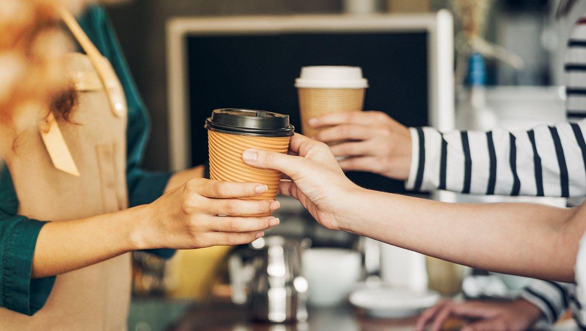 A barista hands a coffee cup to a customer while another person holds a cup in the background.