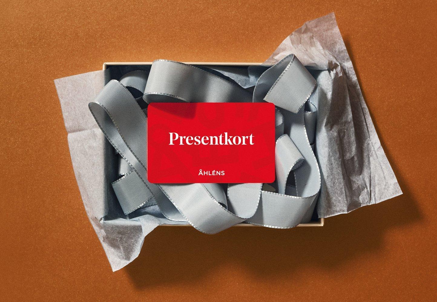 Rött presentkort från Åhléns placerat i en presentask med grått sidenband och silkespapper mot brun bakgrund.